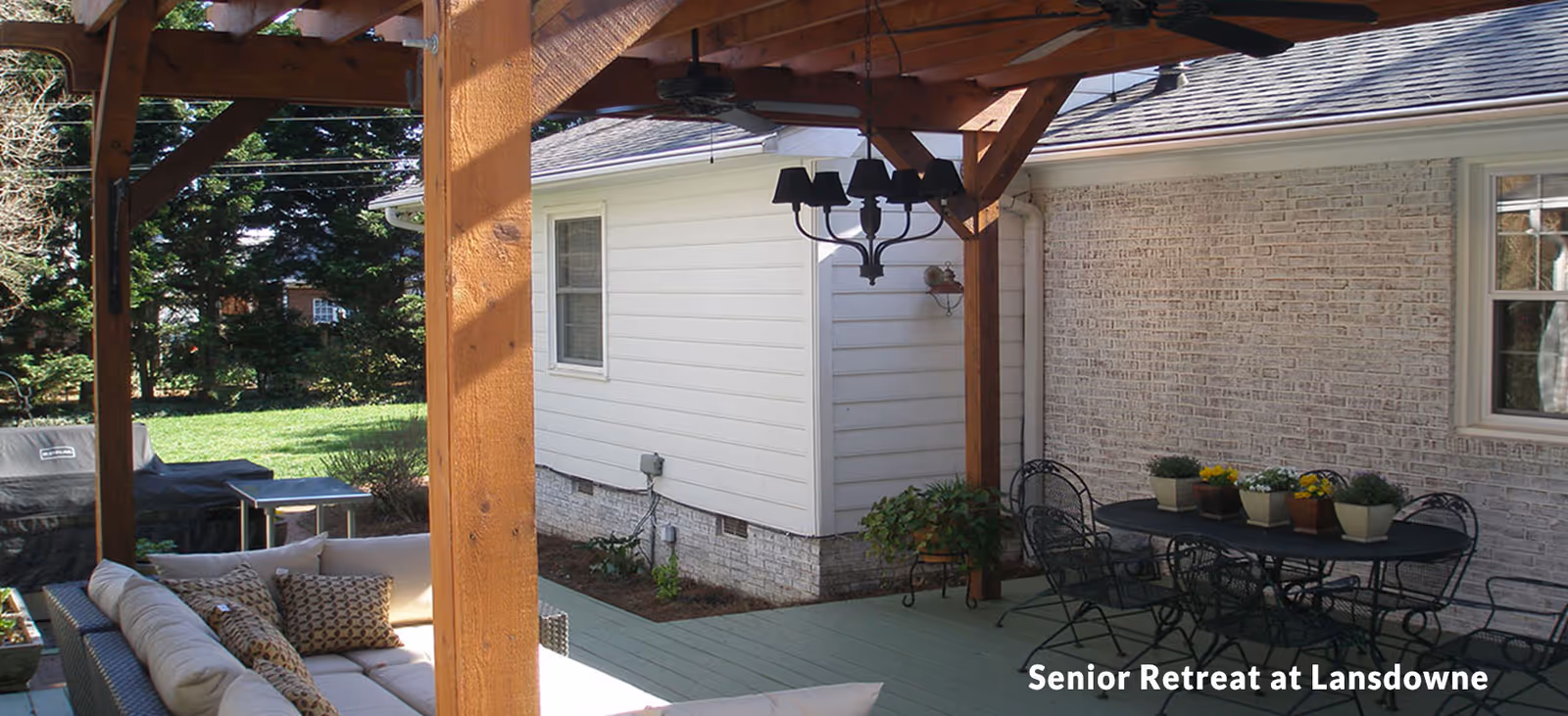 Outdoor patio area with wooden pergola, ceiling fan, and chandelier. There is a cushioned outdoor sofa with patterned pillows on the left and a black metal dining table with chairs and potted plants on the right. The patio is adjacent to a white brick and siding house with a green lawn and trees in the background.