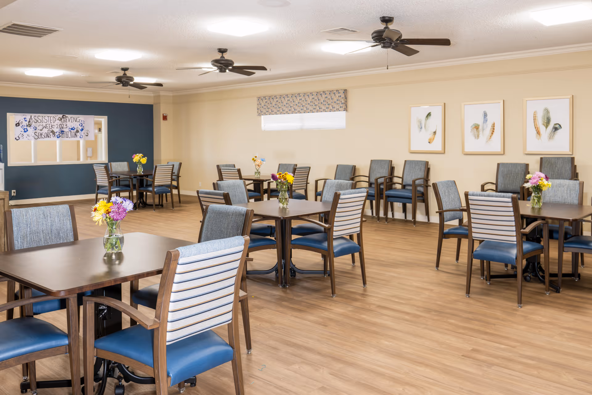 A spacious dining room with multiple wooden tables and chairs arranged neatly. Each table has a small vase with colorful flowers. The room has light-colored walls, three ceiling fans, framed artwork on one wall, and a window with a patterned valance. A banner on a blue accent wall reads 'Assisted Living Week 2023 Season of Reflection.'