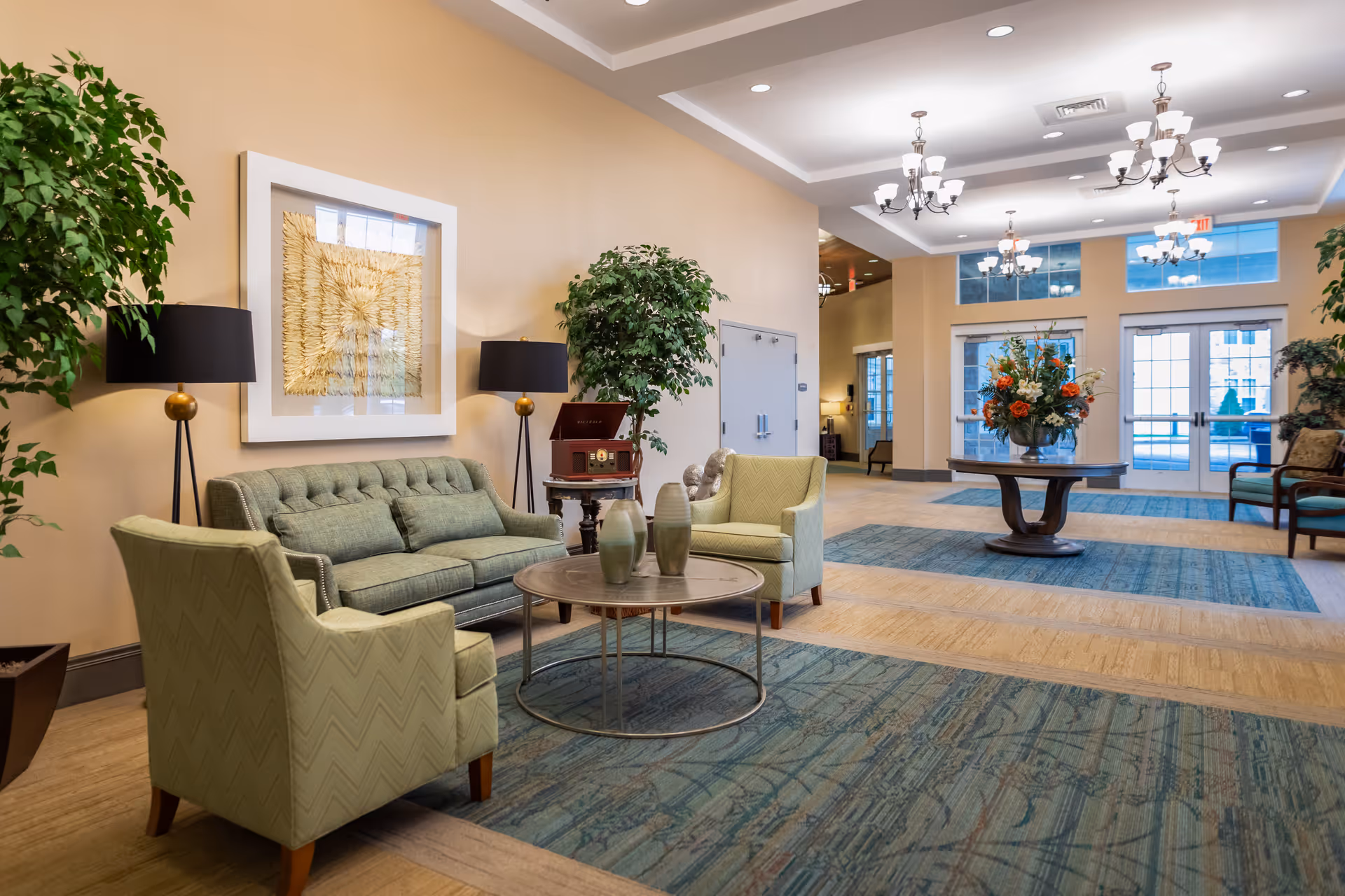 A bright lobby seating area with green sofas and armchairs, coffee tables, plants, chandeliers, and a central floral arrangement by glass doors.