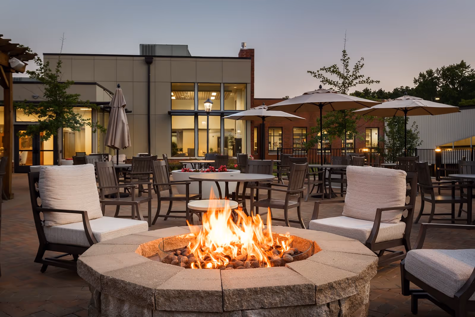 Outdoor patio area at Gambrill Gardens Senior Apartments featuring a stone fire pit with flames, surrounded by cushioned chairs. Several tables with umbrellas and additional chairs are arranged on the brick-paved patio. The building with large windows and trees is visible in the background during dusk.