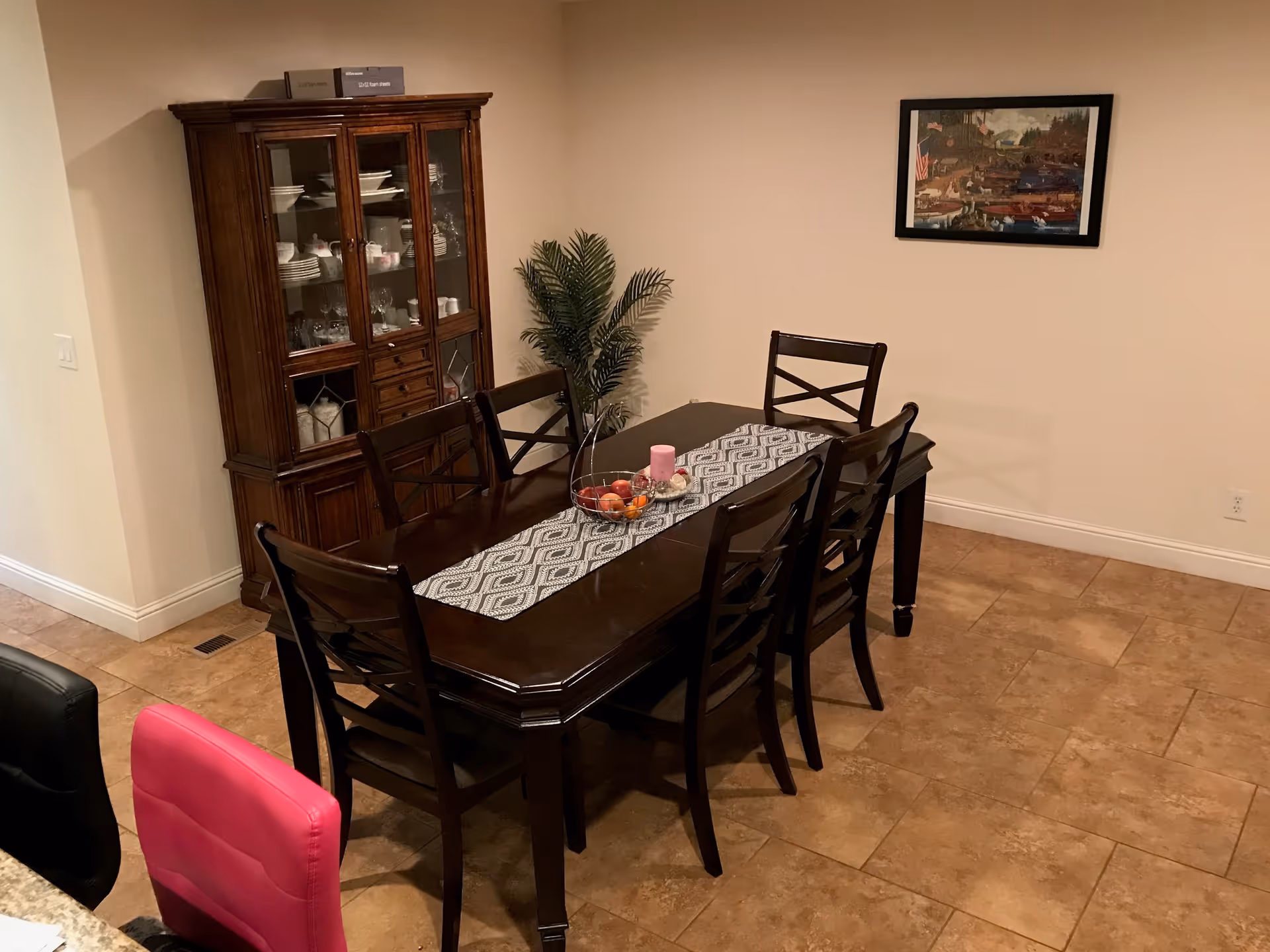 Dining room with a dark wooden table and six matching chairs. A patterned table runner with a bowl of fruit and a pink candle is on the table. A wooden china cabinet filled with dishes is against the wall. There is a framed picture on the wall and a potted plant in the corner. The floor is tiled in a light brown color.