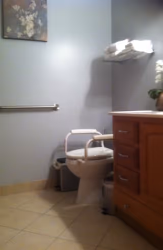A bathroom with a toilet equipped with safety rails, a wooden vanity with drawers, a towel rack holding folded white towels, a small trash can, and a floral wall decoration.