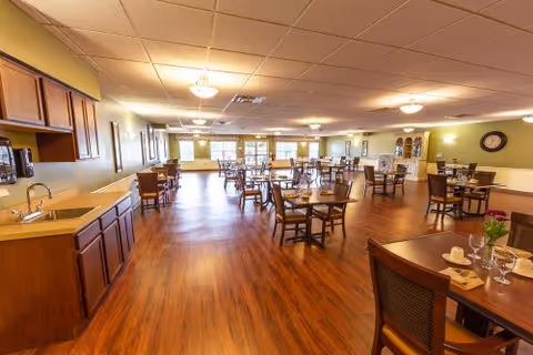 Spacious dining room with wooden floors, multiple tables and chairs arranged neatly, a kitchenette area with cabinets and a sink on the left, and soft lighting from ceiling fixtures.