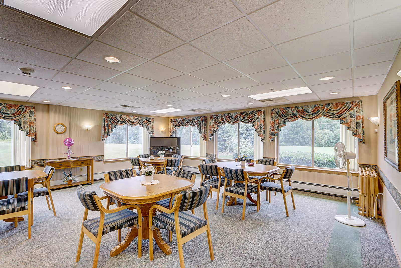 A bright and spacious dining room with multiple wooden tables and striped cushioned chairs arranged neatly. Large windows with floral valances let in natural light and offer a view of greenery outside. The room has a carpeted floor, a standing fan, a clock on the wall, and a TV in the corner.