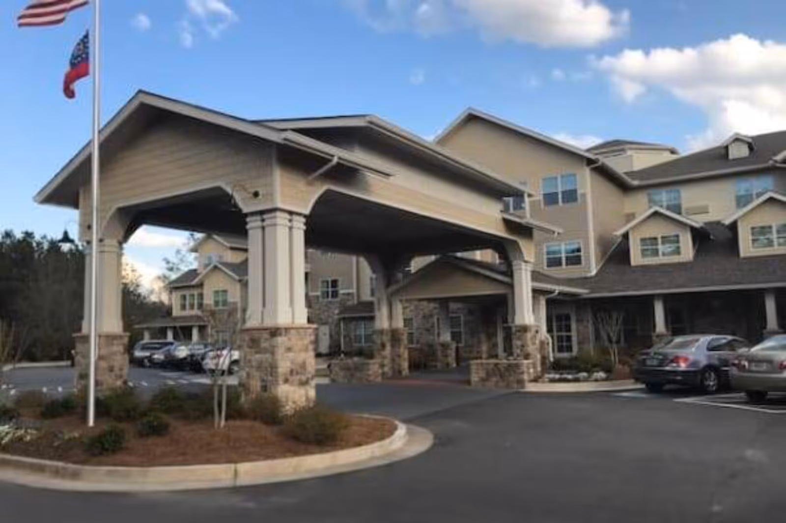 Front entrance and covered porte-cochere of a multi-story senior living facility with flags and parked cars.