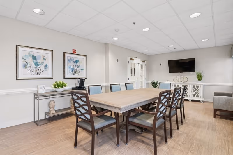 A well-lit meeting or dining room with a large rectangular wooden table surrounded by eight chairs with cushioned seats. The room has light-colored walls adorned with two framed floral paintings. There is a console table with a decorative bust, a bowl of flowers, and a coffee maker. A flat-screen TV is mounted on the wall above a white cabinet with decorative elements and plants. The floor is wooden, and the ceiling has recessed lighting.