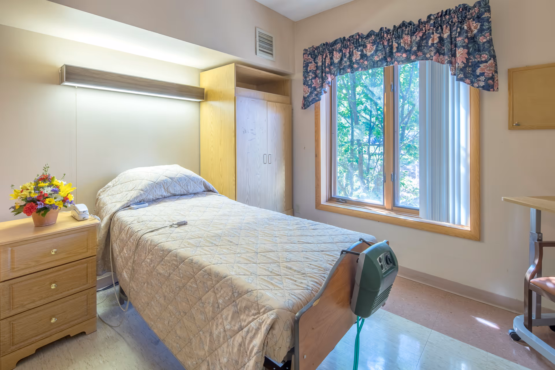 A single bed with a beige quilted bedspread in a nursing facility room. Next to the bed is a wooden nightstand with a colorful flower arrangement and a telephone. Behind the bed is a wooden wardrobe and above the bed is a wall-mounted light fixture. A window with floral curtains lets in natural light, showing green trees outside. The room has light-colored walls and flooring.