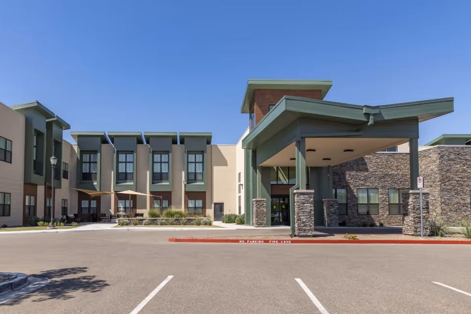 Exterior view of Surprise Health & Rehabilitation Center showing the building entrance with a covered drop-off area supported by stone pillars, multiple windows, and a clear blue sky above. There is a parking area in front with marked spaces and a red curb labeled 'No Parking Fire Lane'.
