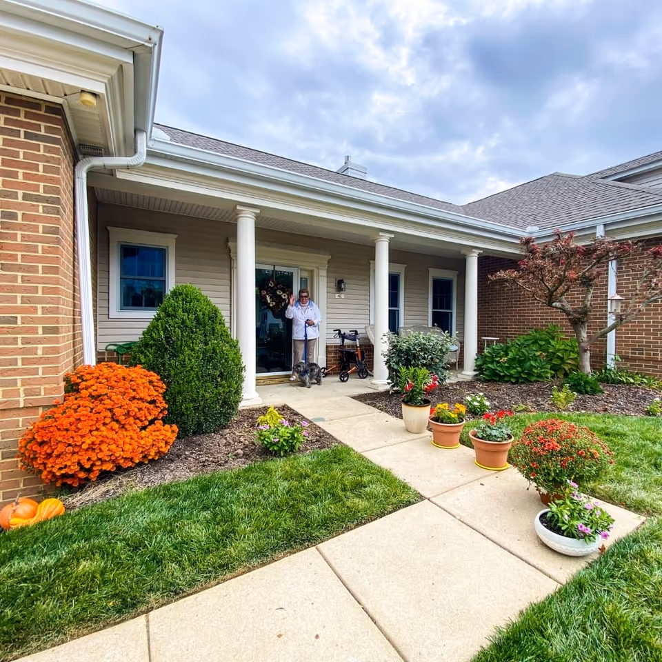 Front porch of a residential unit at The Knolls of Oxford, a CCRC, featuring a person standing with a small dog near the entrance. The porch has white columns, a wreath on the door, and a walker nearby. The surrounding garden has green grass, orange flowers, potted plants, and a small tree under a cloudy sky.