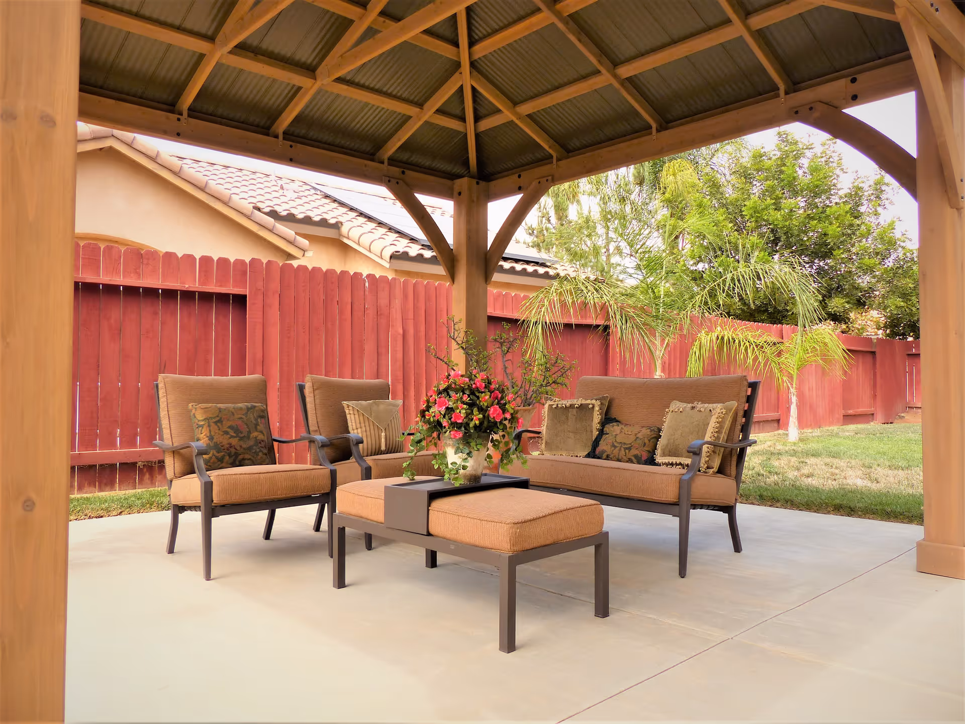 Outdoor seating area under a wooden gazebo with cushioned chairs, a loveseat, and an ottoman with a flower arrangement on top. The area is surrounded by a red wooden fence and greenery including palm trees and other plants.