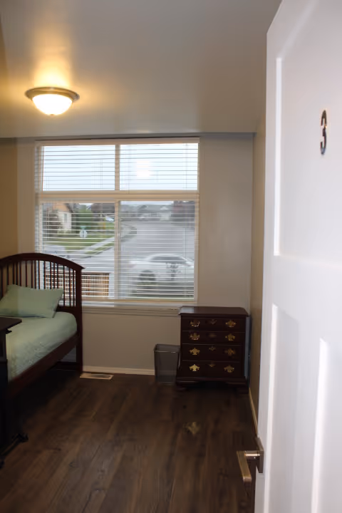 View into a small bedroom with a wooden bed frame and green bedding on the left side, a window with blinds showing a street and houses outside, a small wooden dresser with brass handles, and a white door with the number 3 partially open on the right side.