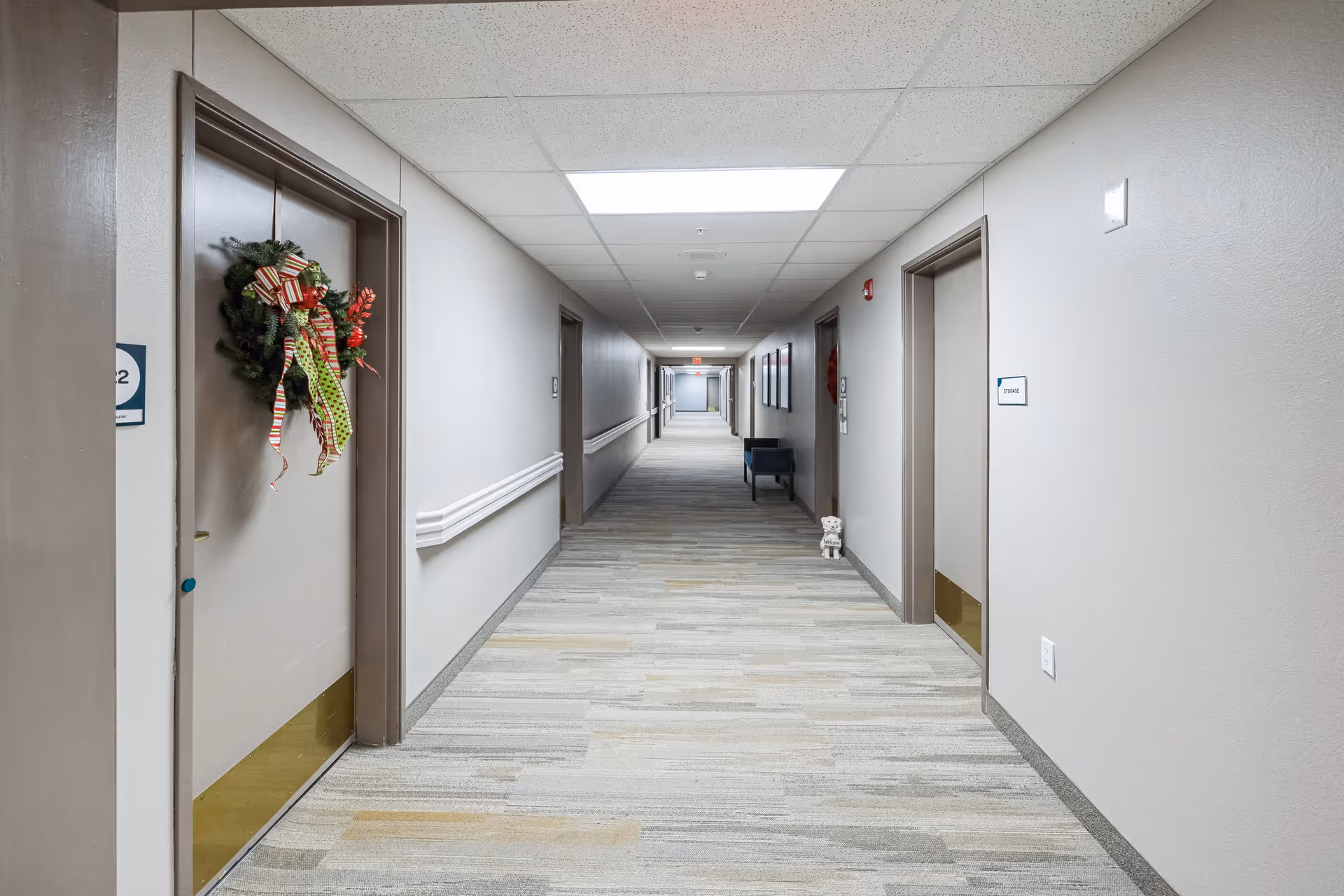 A long, well-lit hallway in a senior living facility with beige walls and carpeted floor. Several doors line both sides of the hallway, one decorated with a holiday wreath. Handrails run along the walls, and a small decorative dog figurine is placed near one door. The ceiling has recessed lighting panels.