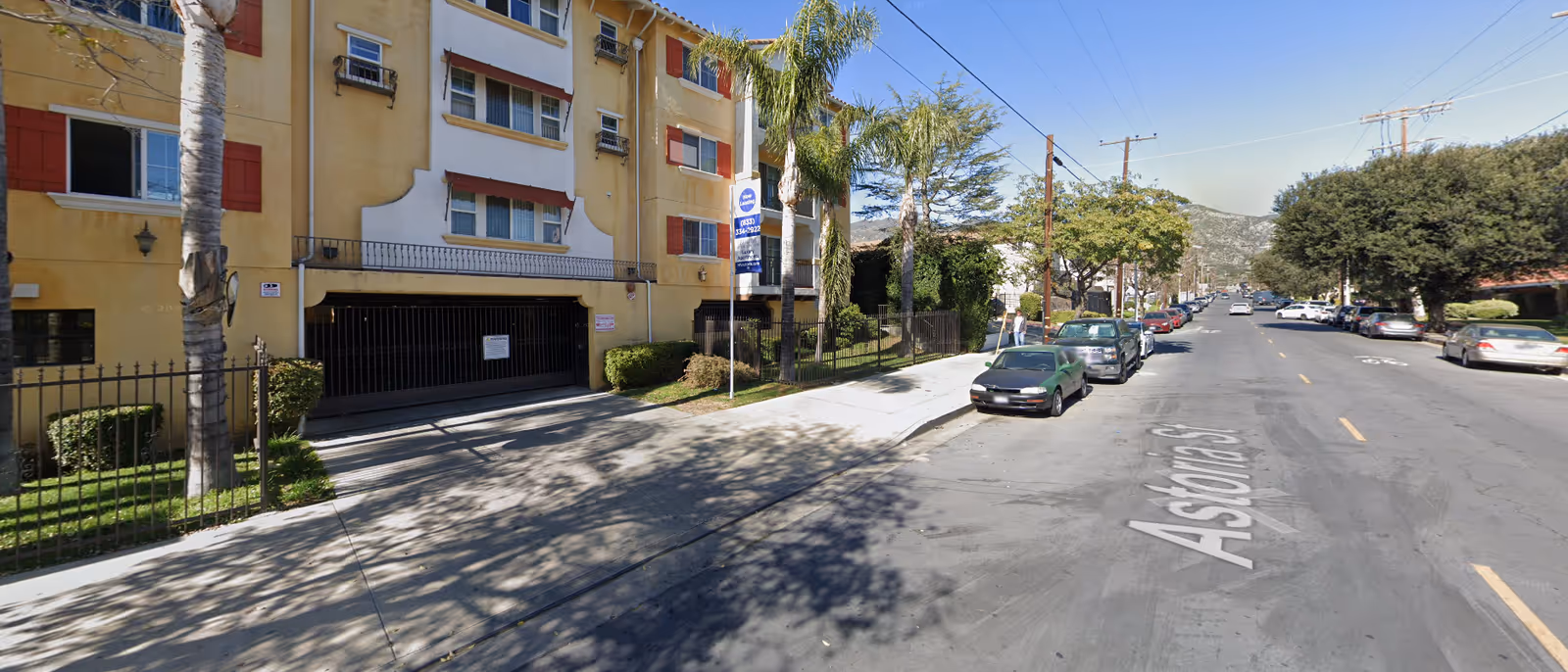 Street view of a residential building with yellow and white exterior walls, red window shutters, palm trees, and parked cars along the street named Astoria St under a clear blue sky.