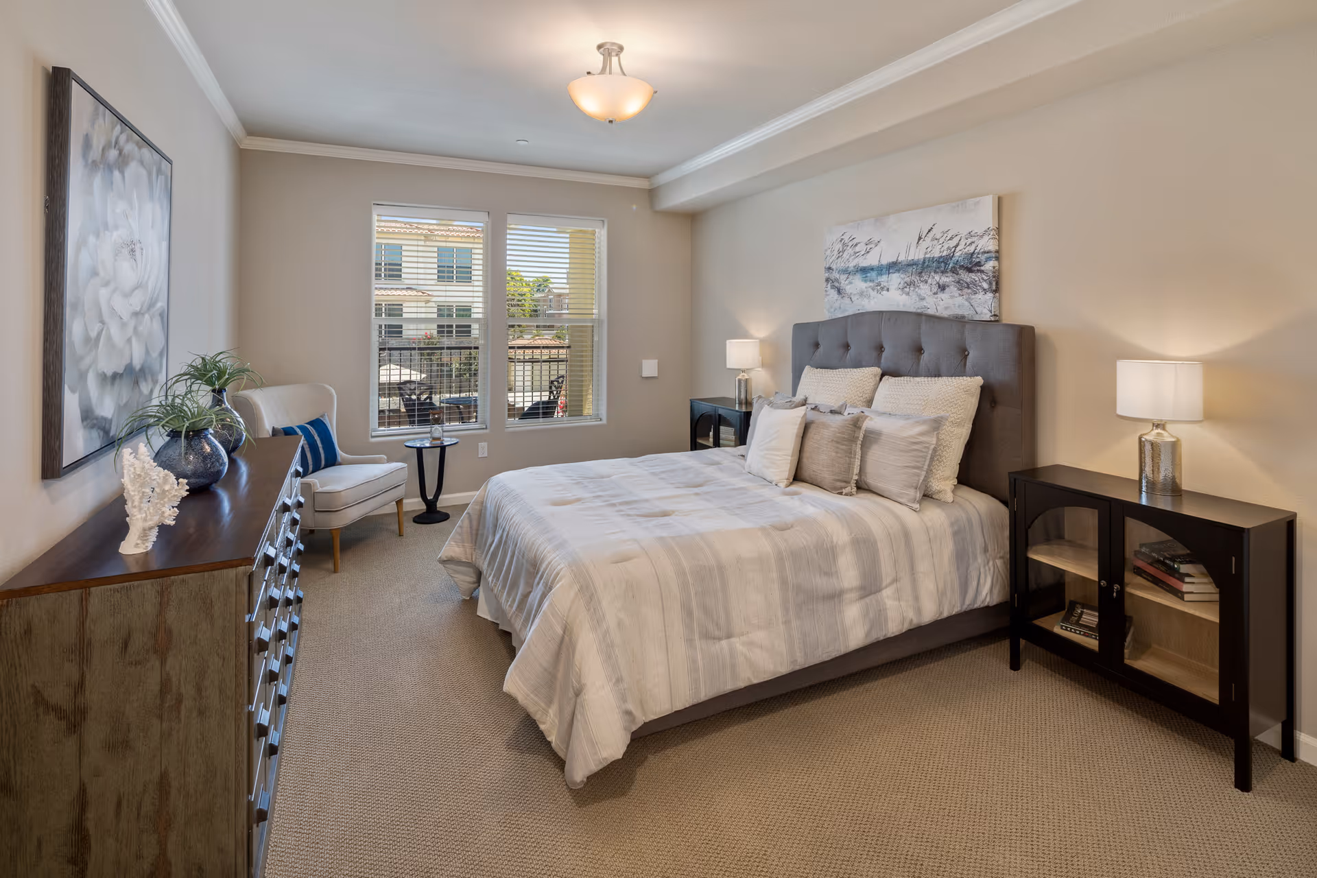 A well-lit bedroom with a large bed featuring a gray upholstered headboard and multiple pillows. On the right side of the bed is a black cabinet with glass doors and a lamp on top. On the left side of the room is a wooden dresser with decorative items and a white armchair next to a small round table. Two windows with blinds allow natural light to enter, and a large floral painting hangs on the left wall while a beach-themed painting is above the bed.