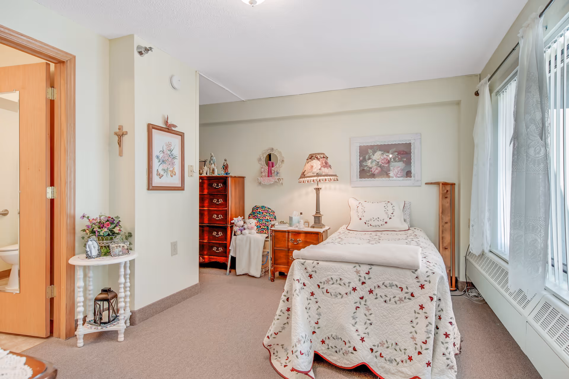 A cozy bedroom in an assisted living facility featuring a single bed with a floral quilt and pillow, a wooden nightstand with a decorative lamp, a wooden dresser, a chair with a colorful blanket, and a small round table with flowers and framed photos. The room has large windows with sheer white curtains allowing natural light to fill the space. A bathroom door is open, showing a glimpse of the toilet inside. The walls are decorated with framed floral artwork and a small crucifix.