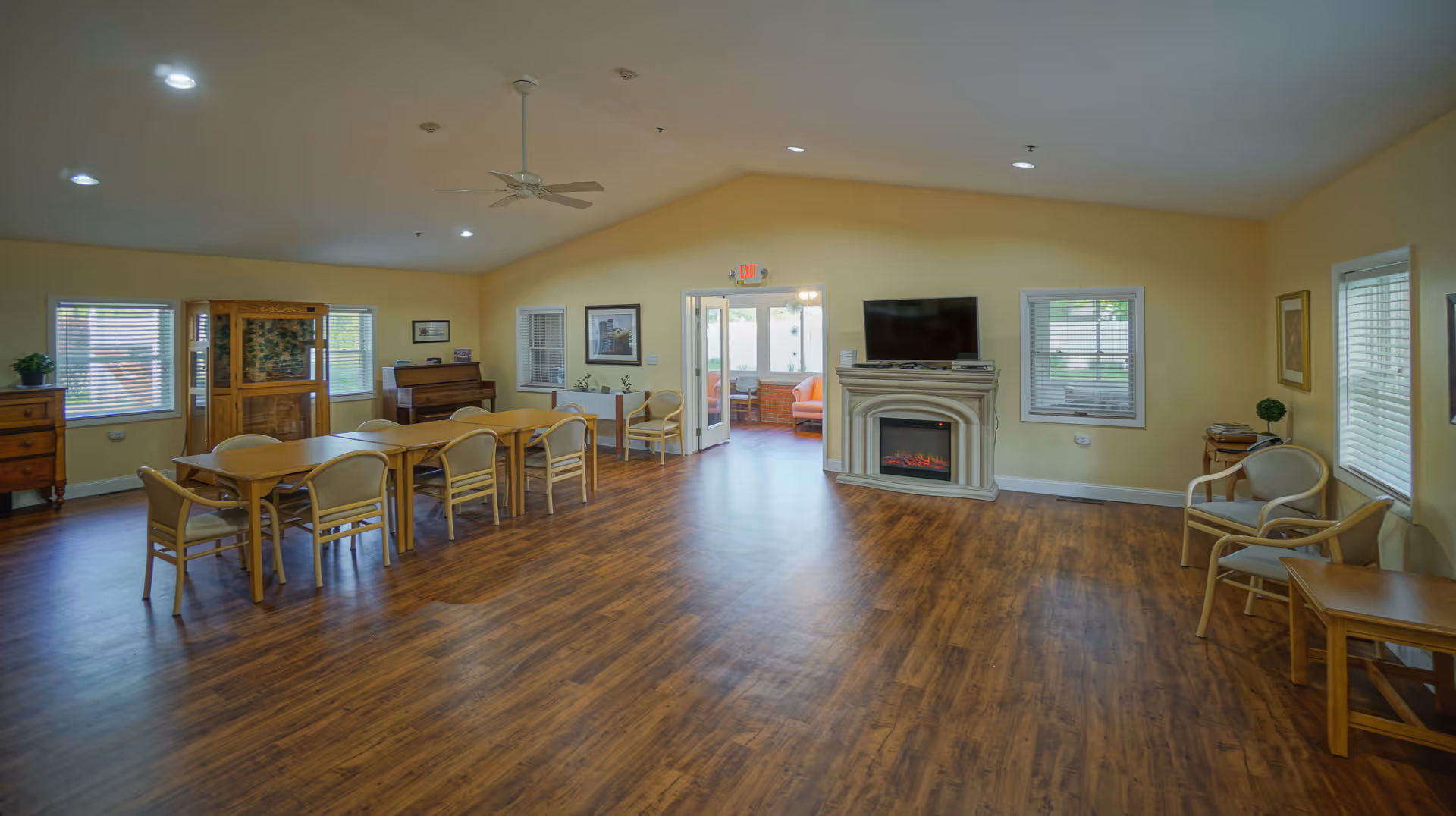 Spacious common area with wooden flooring, several chairs and tables, a piano, a fireplace with a TV mounted above it, and multiple windows letting in natural light. The walls are painted a soft yellow, and there is a ceiling fan and recessed lighting on the ceiling.