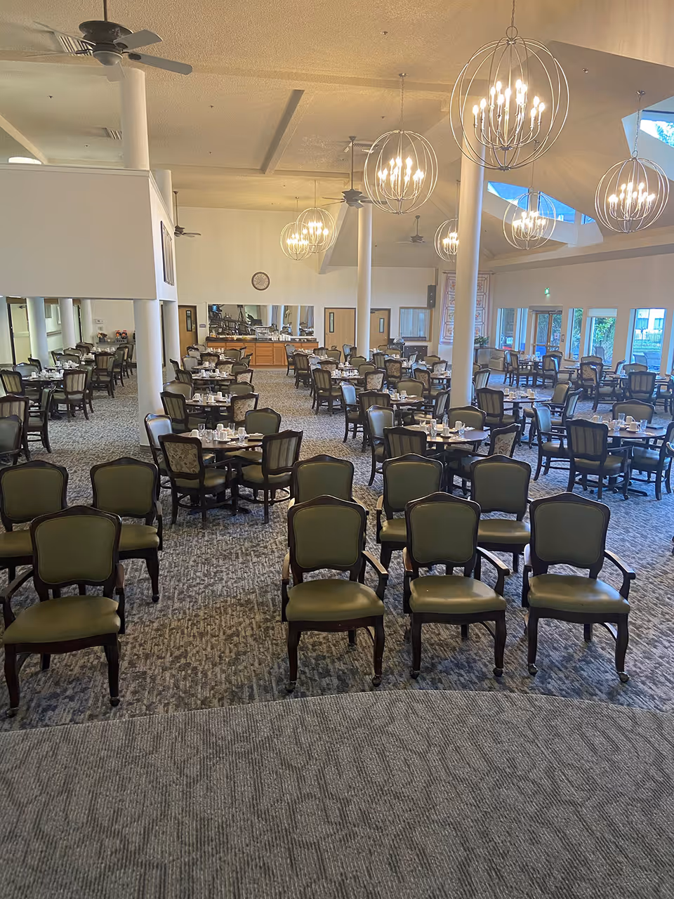 Empty dining room with many round tables set for service and rows of green upholstered chairs beneath modern chandeliers.