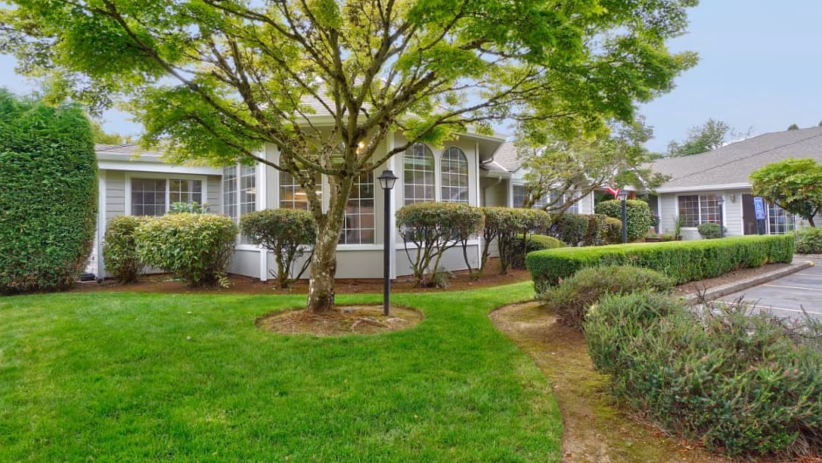 Front exterior of a single-story senior living building with large windows, trimmed hedges, a tree, and a grassy lawn.