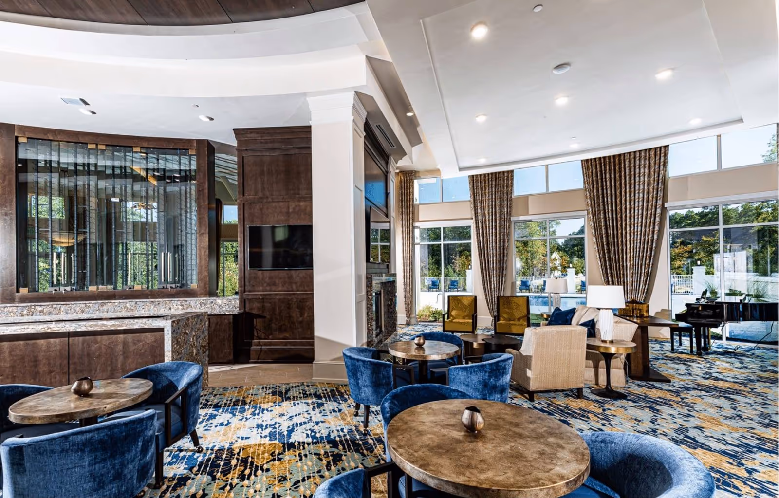 A spacious and well-lit living room area with large windows and tall curtains allowing natural light to fill the space. The room features multiple round tables with blue upholstered chairs, a seating area with beige armchairs and yellow accent chairs, a fireplace with a mounted TV above it, and a grand piano near the windows. The carpet has a colorful abstract pattern in blue, yellow, and beige tones.