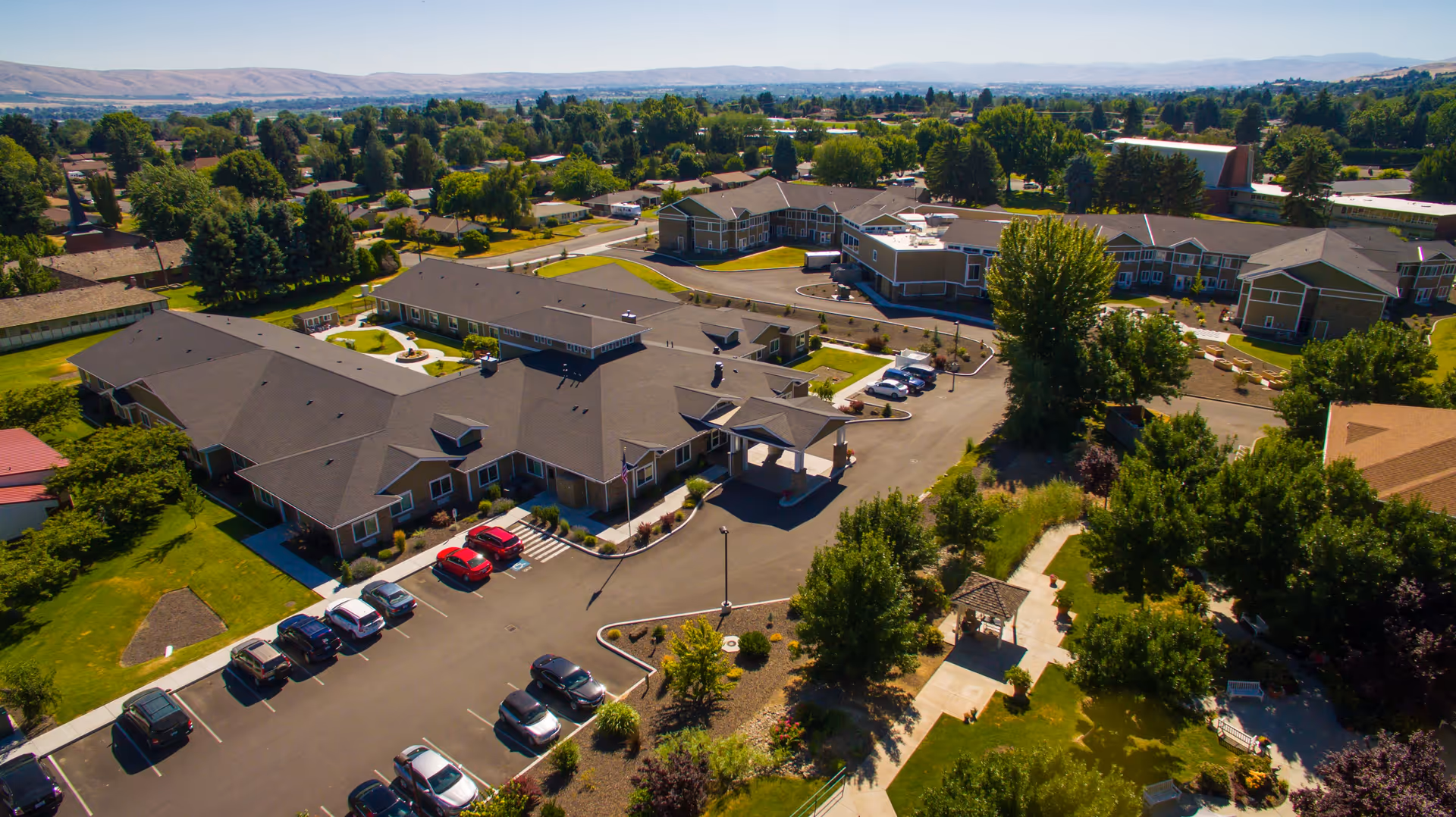 Aerial view of Fieldstone Memory Care Yakima facility showing multiple connected buildings with parking lots, surrounded by trees and greenery, with distant hills in the background under a clear sky.