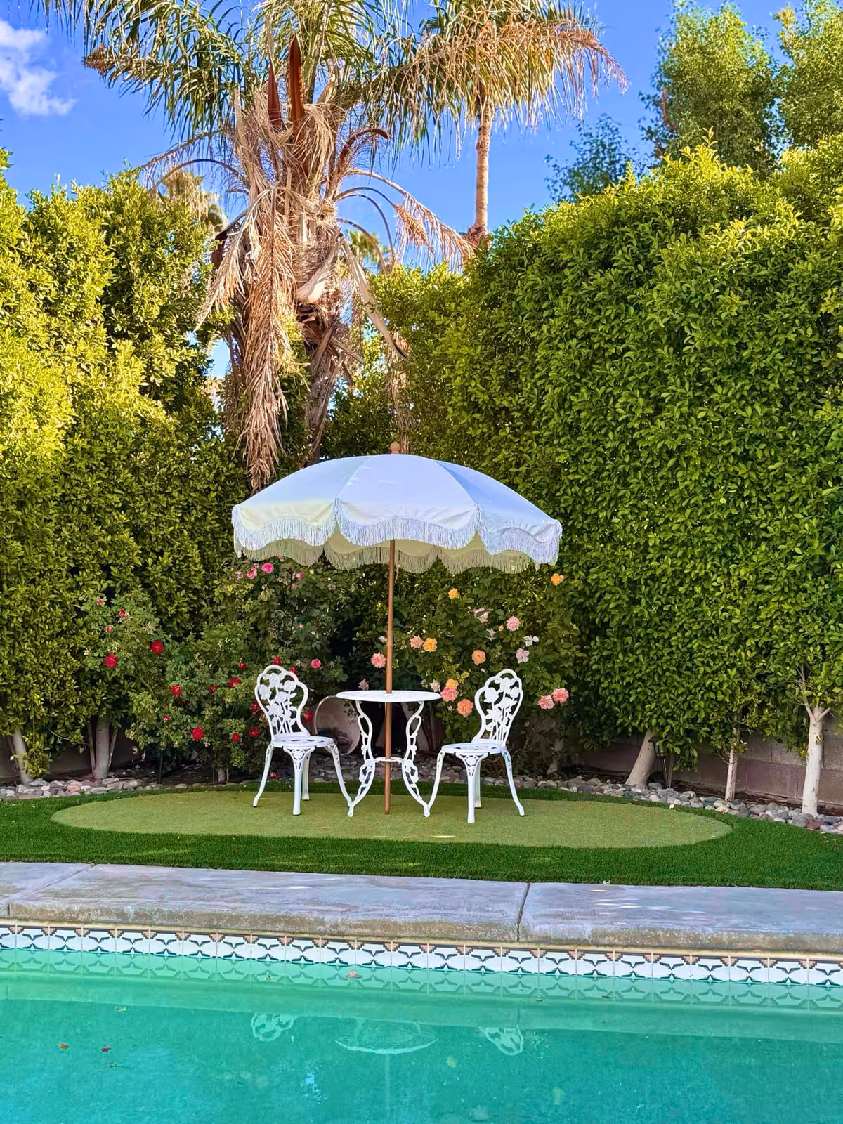 A small outdoor seating area with two white ornate metal chairs and a round white table under a white umbrella with fringe, set on a patch of green artificial grass next to a swimming pool. The background features tall green bushes, flowering plants, and palm trees under a clear blue sky.