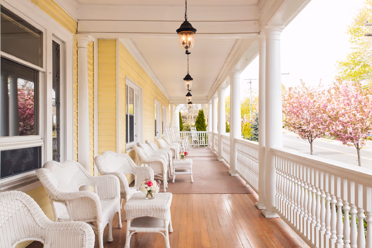 Covered front porch with white wicker chairs and small tables along a yellow-sided building, white columns, and blooming trees beyond.