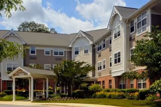 Exterior view of a multi-story senior living facility building with beige and white siding, multiple windows, and a covered entrance with a bench. The surrounding area has well-maintained landscaping with trees, bushes, and a lawn under a partly cloudy sky.