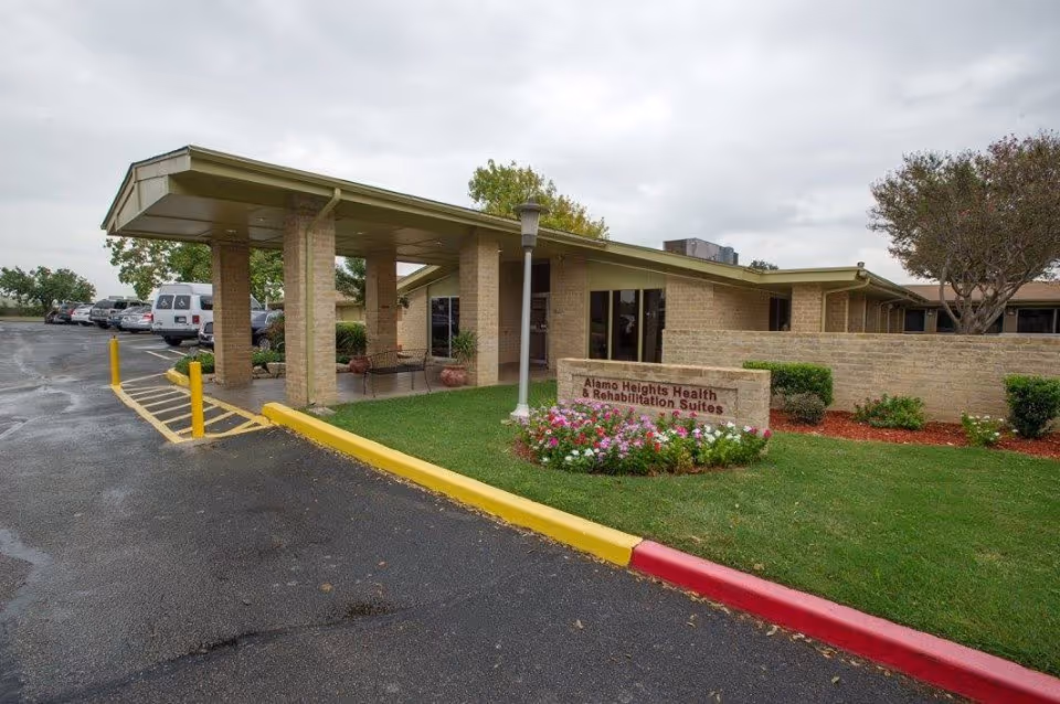 Front entrance and covered drop-off (porte-cochère) of a single-story rehabilitation facility with a sign, parking area, and landscaped flowerbeds.