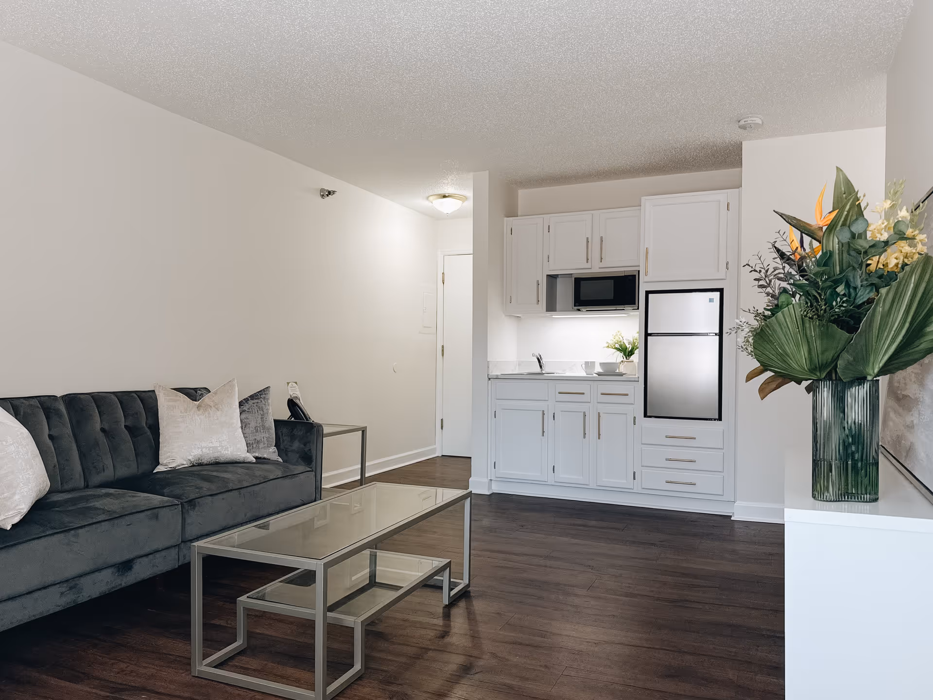 A modern living area with a dark gray velvet sofa adorned with light-colored pillows, a glass coffee table with a smaller nested table underneath, and a small kitchenette with white cabinets, a microwave, and a mini refrigerator. There is a large vase with green and yellow flowers on a white cabinet to the right, and the floor is dark wood.
