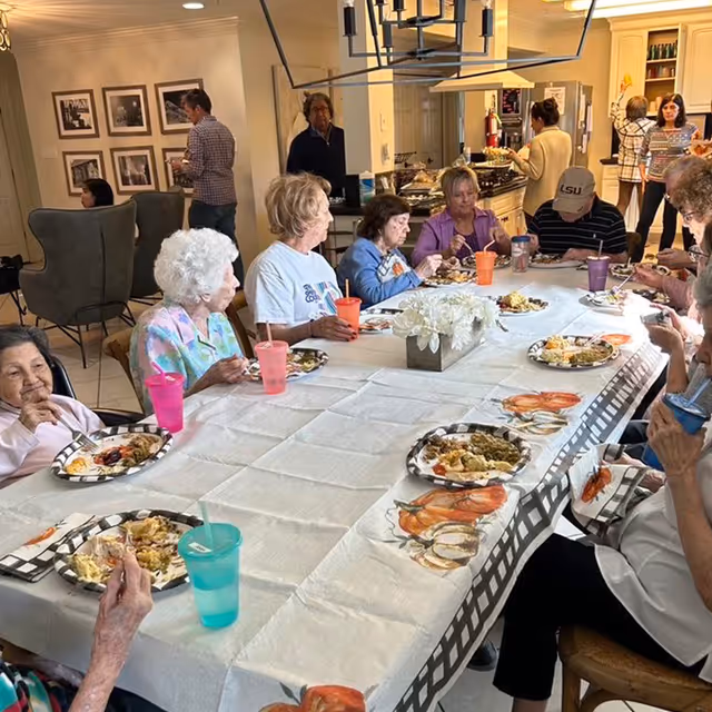 Several elderly residents seated around a long table in a communal dining area enjoying a meal.