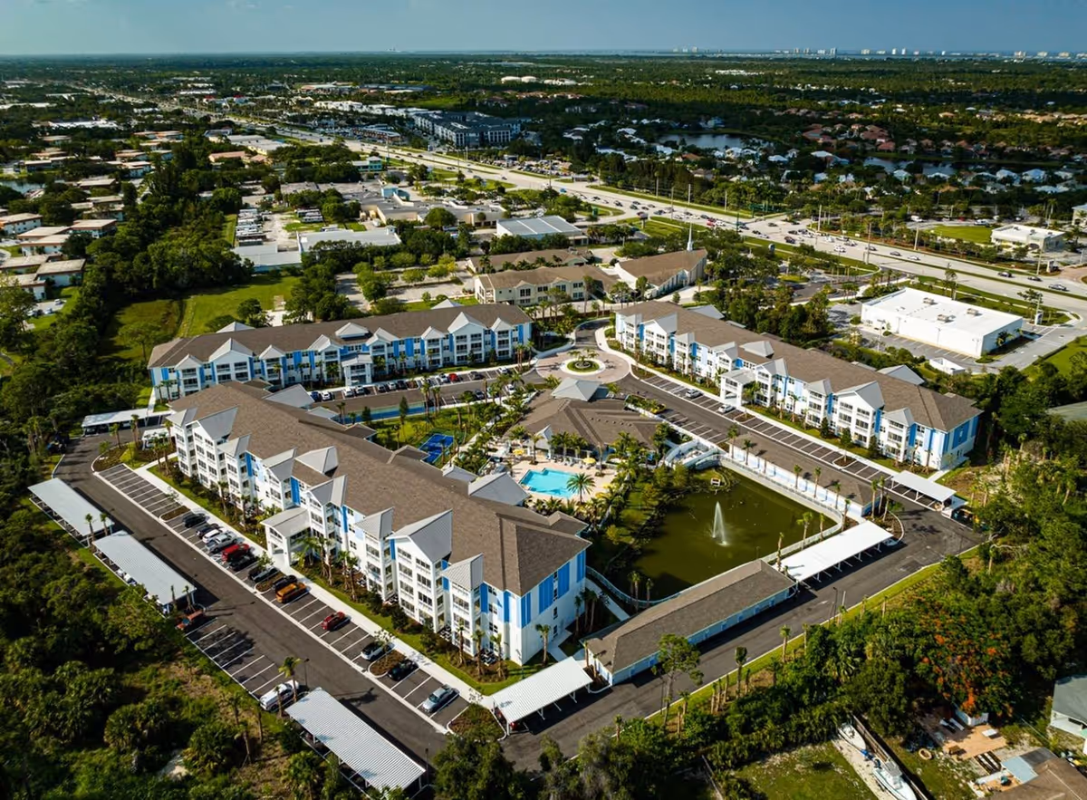Aerial view of The Sovana At Stuart senior living facility showing multiple connected buildings with brown roofs and white and blue exterior walls. The facility includes a central courtyard with a swimming pool, a pond with a fountain, palm trees, and surrounding parking lots. The surrounding area features roads, greenery, and other buildings in the distance.