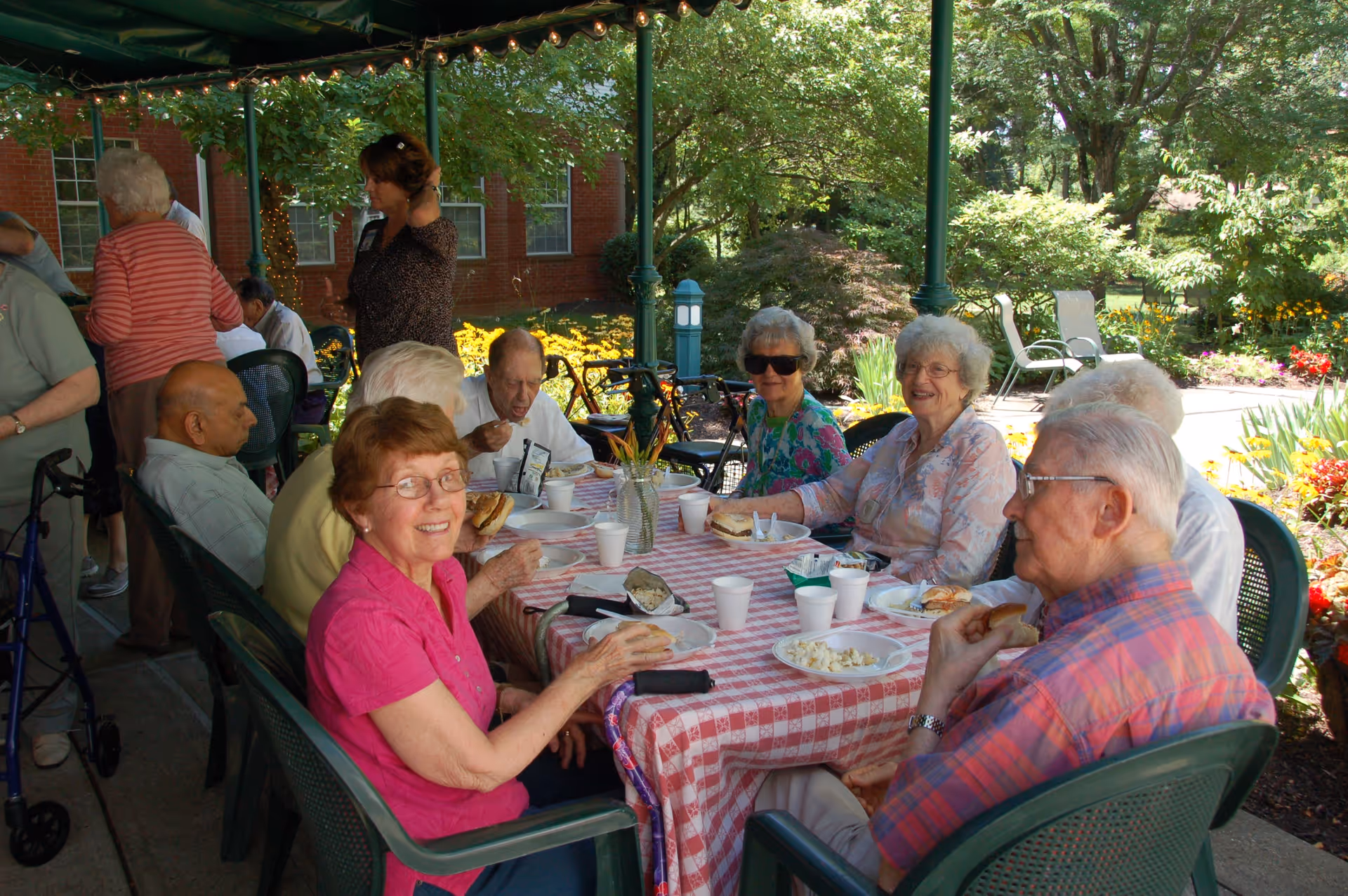 A group of elderly people sitting around a table outdoors under a canopy, enjoying a meal together. The table is covered with a red and white checkered tablecloth and has plates of food, cups, and a vase with flowers. The background shows greenery, flowers, and part of a brick building.