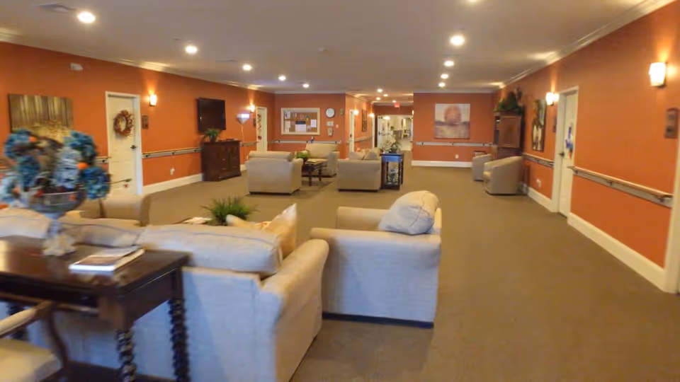 A spacious common area with beige armchairs and sofas arranged in small groupings. The walls are painted a warm orange color with white trim, decorated with framed artwork and wall sconces. There is a wooden table with a floral arrangement and books in the foreground. The room is well-lit with recessed ceiling lights and has a carpeted floor.