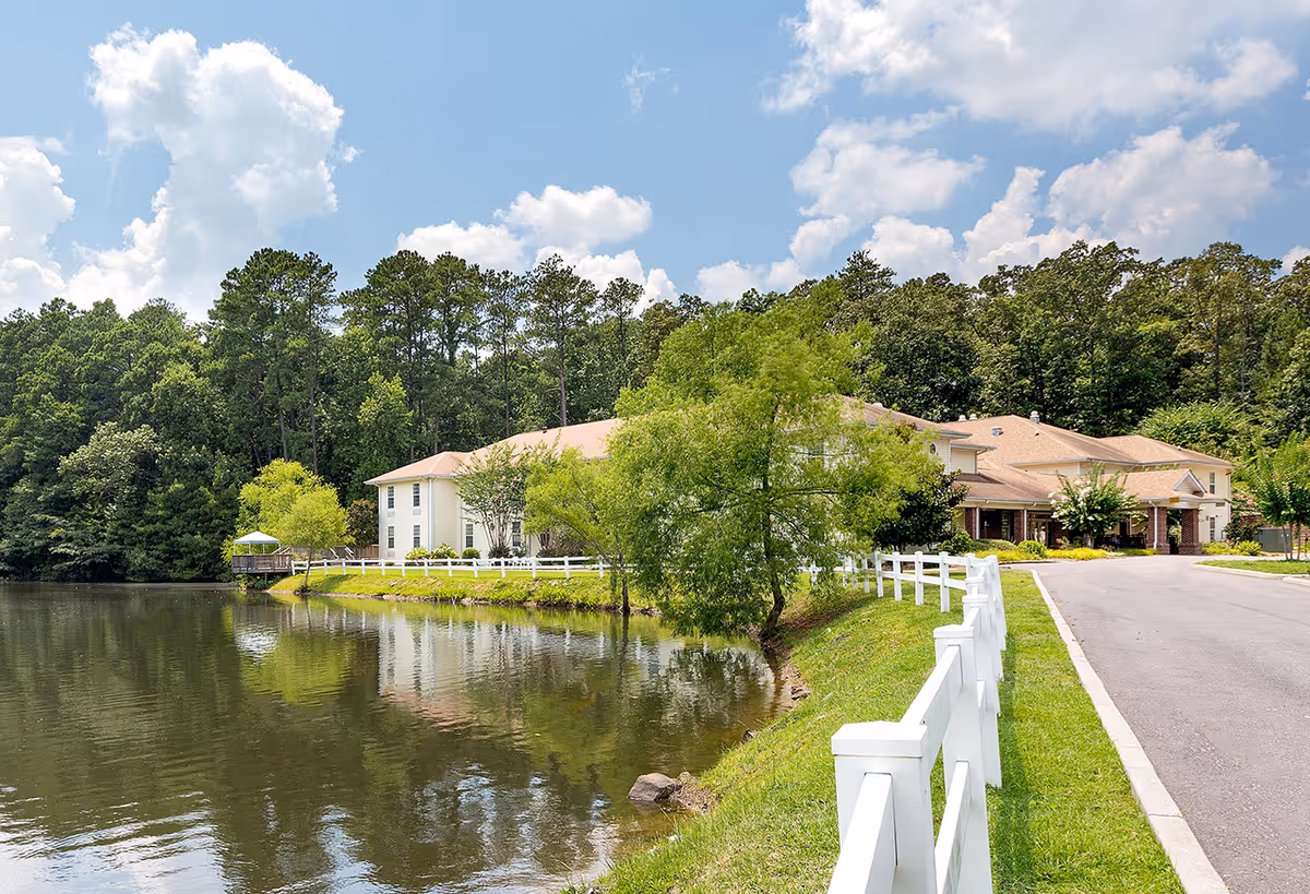 Exterior view of a senior living facility named Lakeview Estates - Memory Care Community, showing a large building with a beige roof surrounded by trees and greenery. There is a white fence along a paved driveway and a calm lake reflecting the building and trees under a partly cloudy sky.
