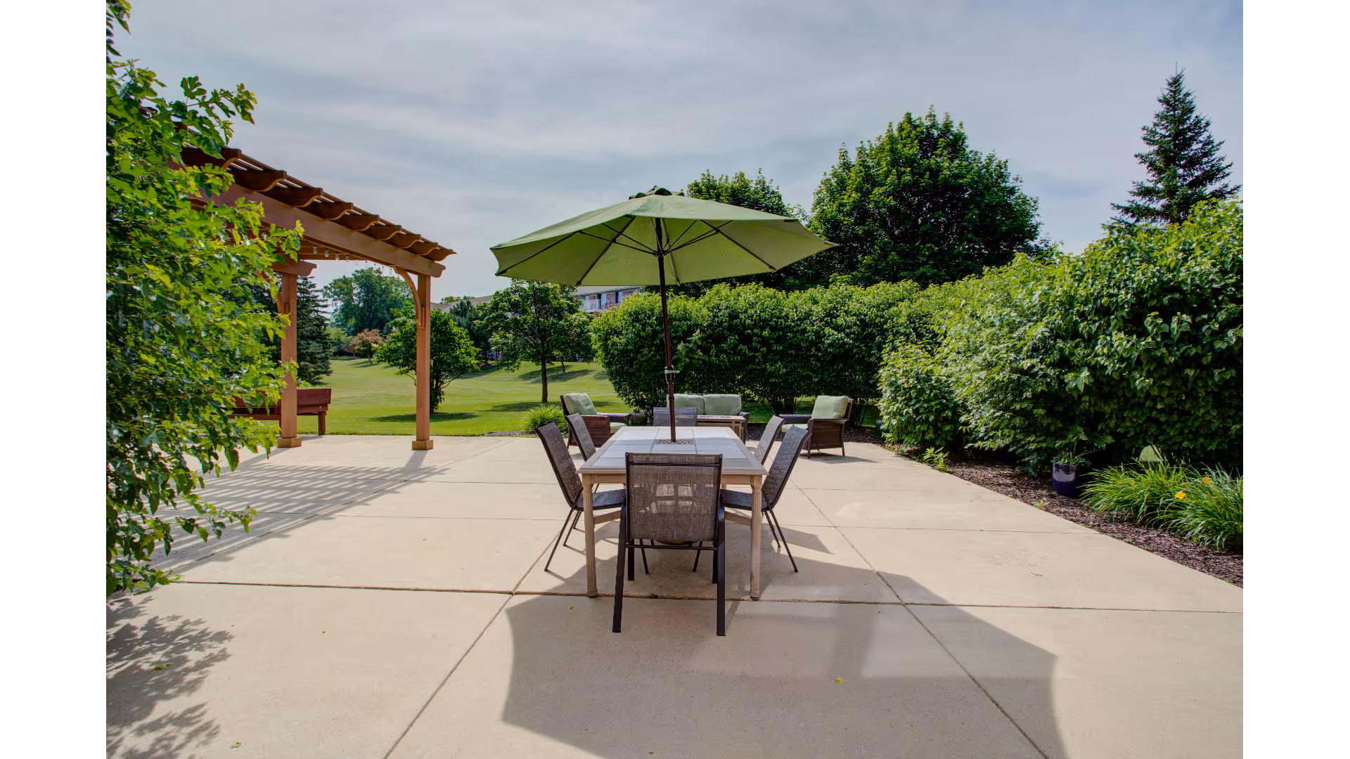 Outdoor patio with a table, chairs and a green umbrella beside a pergola overlooking landscaped lawns and trees.
