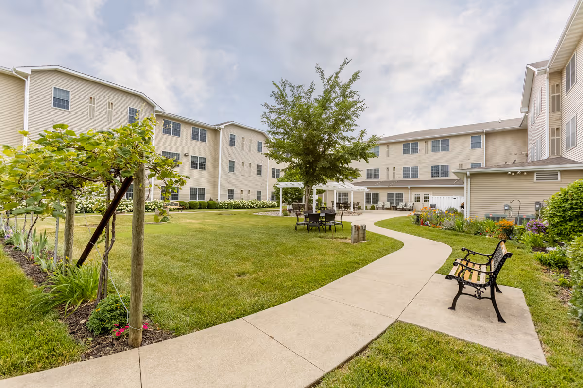 Outdoor courtyard area at Independence Village of Pella featuring a curved concrete walkway, green lawn, garden beds with flowers, a tree with a pergola and outdoor seating, and a bench along the path. The courtyard is surrounded by a three-story beige residential building under a partly cloudy sky.