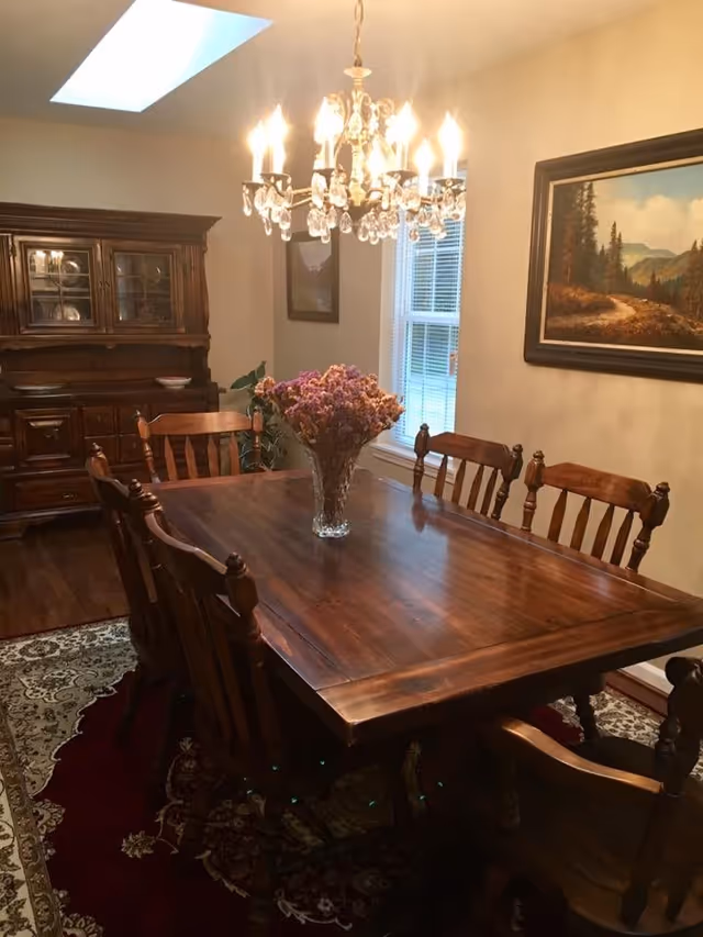 A traditional dining room with a large wooden table surrounded by six matching wooden chairs. A crystal chandelier hangs above the table, which has a vase of dried flowers as a centerpiece. The room features a window with blinds, a wooden china cabinet, a framed landscape painting on the wall, and a skylight providing natural light. A patterned rug is underneath the table and chairs.