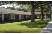 Single-story brick building with multiple windows and white trim, surrounded by green grass and large trees, with a concrete sidewalk running alongside the building.
