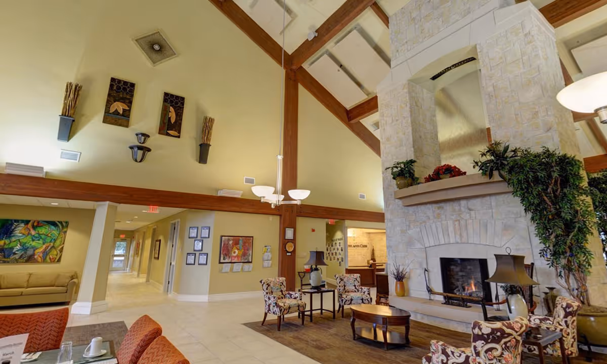 A spacious senior living facility common area with high vaulted ceilings supported by wooden beams. The room features a large stone fireplace with plants on the mantel, several patterned armchairs around a wooden coffee table, and a beige sofa against the far wall. The walls are decorated with colorful artwork and wall-mounted sconces. There is a hallway leading to other rooms and a reception desk visible in the background.