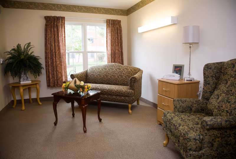 Cozy living room with a patterned loveseat and armchair, wooden coffee table with flowers, side tables, lamp, and a window with curtains.