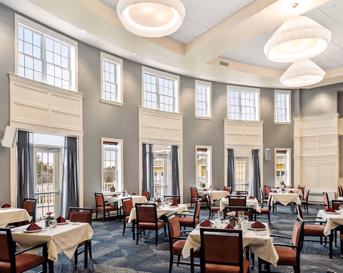 A spacious dining room with multiple tables covered in beige tablecloths, each set with folded napkins, glasses, and silverware. The room features tall windows with white trim and dark curtains, high ceilings with large modern pendant lights, and a patterned blue carpet.