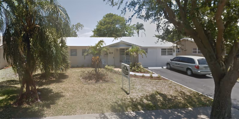 Single-story building with a white roof and beige walls, surrounded by palm trees and other greenery. A driveway with a parked silver minivan is visible on the right side. A sign is placed on the lawn in front of the building.