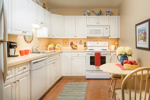 A bright kitchen with white cabinets and appliances including a stove, microwave, dishwasher, and refrigerator. The countertop has decorative items like ceramic jars and a rooster figurine. A small round dining table with two chairs is set with a basket of apples and a vase of white flowers. A framed picture hangs on the wall, and a blue rug lies on the wooden floor.