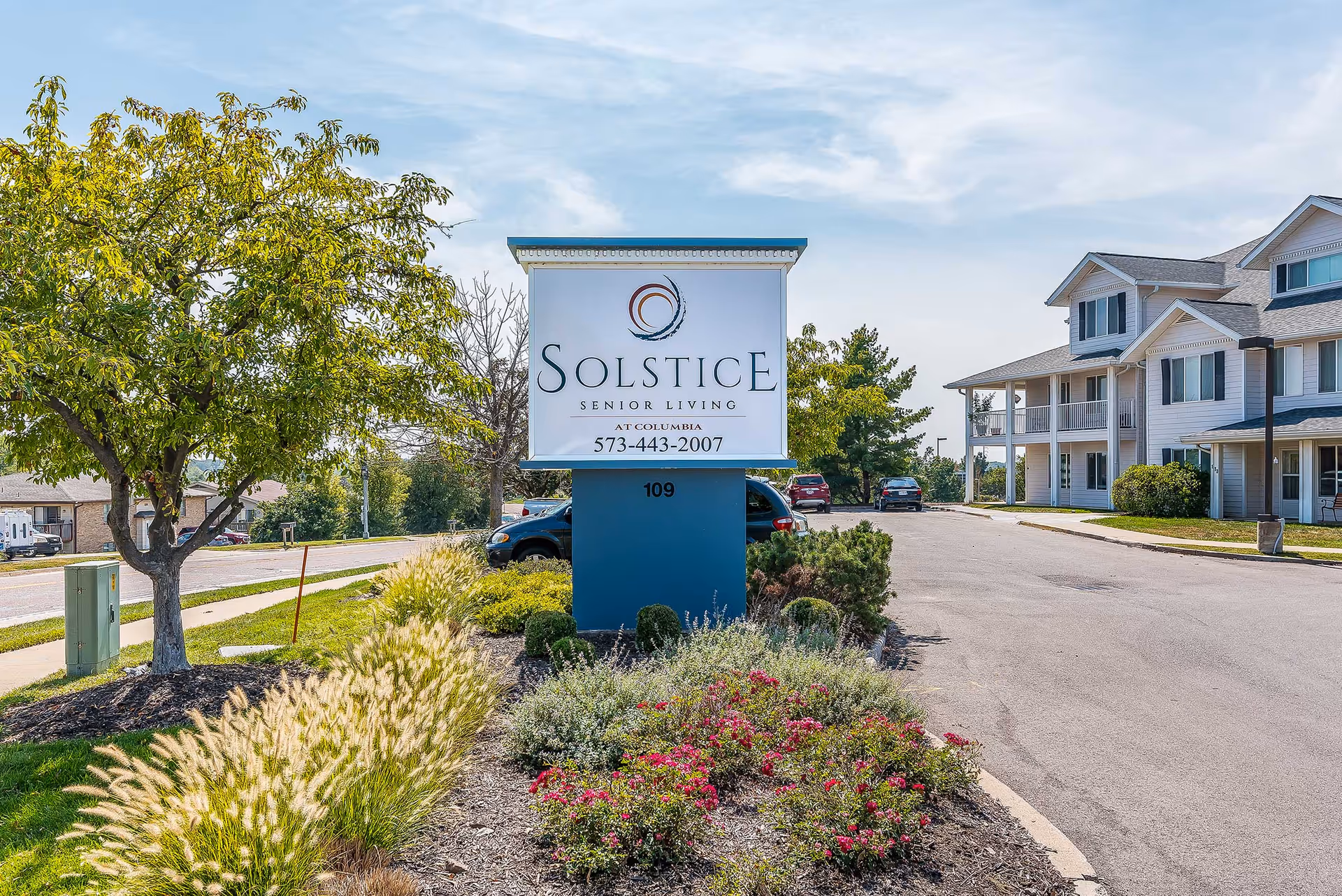 Outdoor view of Solstice Senior Living at Columbia facility sign with landscaping including bushes, flowers, and a tree. A paved driveway and part of the senior living building with white siding and balconies are visible in the background under a partly cloudy sky.