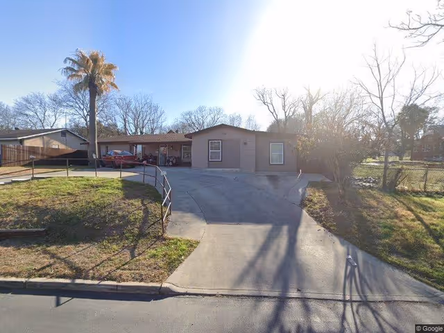 Front exterior of a single-story house with a driveway, carport, parked cars, and a tall palm tree.