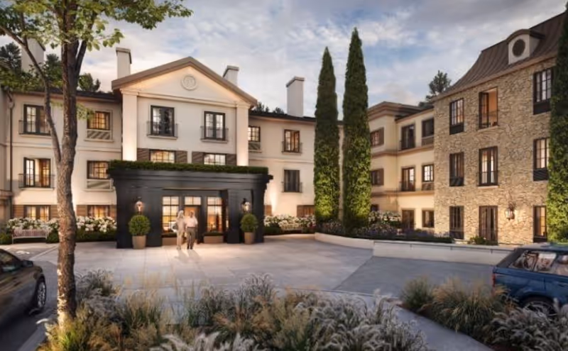Front entrance of a three-story Mediterranean-style senior living building with landscaped planters, parked cars, and two people near the main doors.
