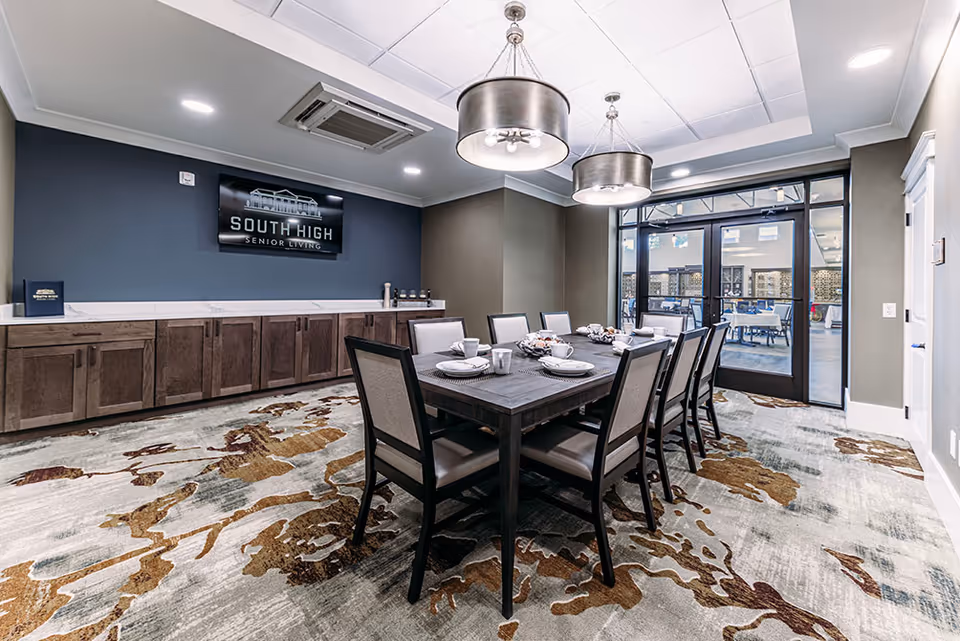 A modern dining room in South High Senior Living featuring a large rectangular wooden table set for eight with plates, cups, and napkins. The room has a patterned carpet, two large hanging light fixtures, a dark blue accent wall with a mounted TV displaying the South High Senior Living logo, and a long wooden cabinet beneath the TV. Glass doors lead to an outdoor seating area.