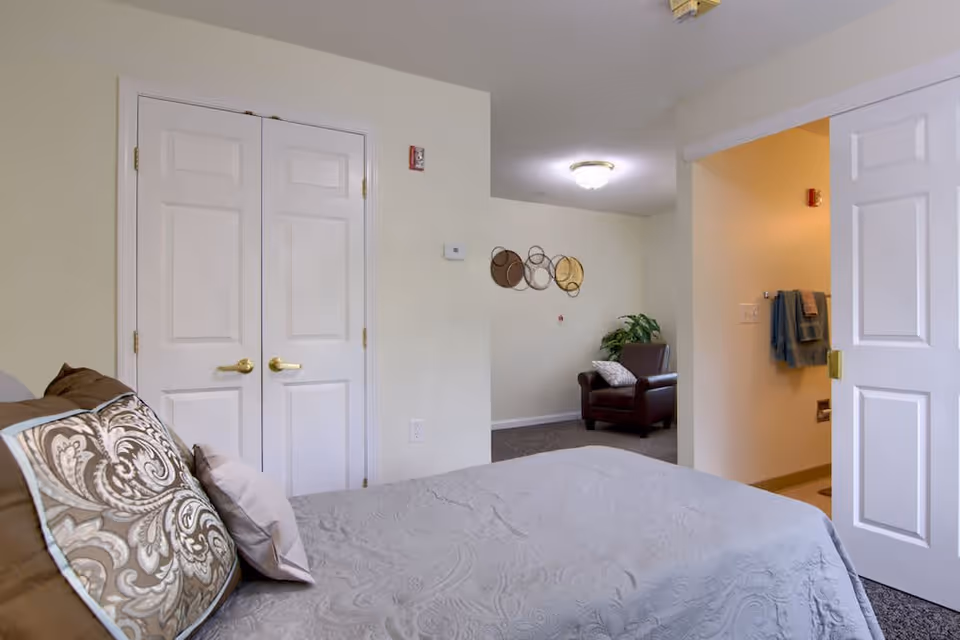 A bedroom in a senior living facility featuring a bed with patterned pillows and a light gray bedspread. There is a double-door closet with gold handles on the left wall. An open doorway on the right leads to a bathroom with towels hanging on the wall. In the background, a brown armchair with a pillow is placed against a wall decorated with circular wall art. The walls are painted light beige and the room is softly lit.