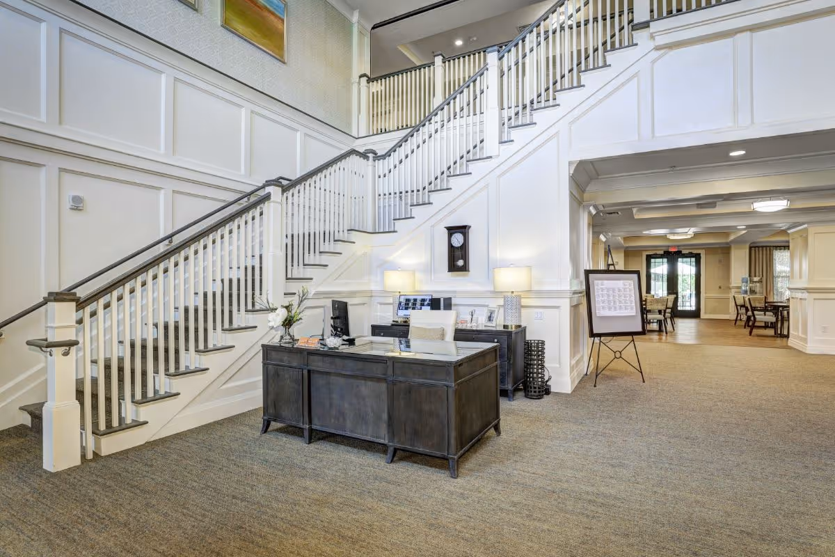 Interior view of a senior living facility lobby with a large staircase, a dark wooden reception desk with a computer and lamps, a wall clock, and a hallway leading to a dining area with tables and chairs.