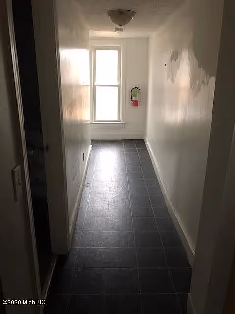 A narrow hallway with dark tiled flooring and white walls, illuminated by natural light coming through a window at the end. A fire extinguisher is mounted on the wall near the window. There is a ceiling light fixture and a partially open door on the left side.